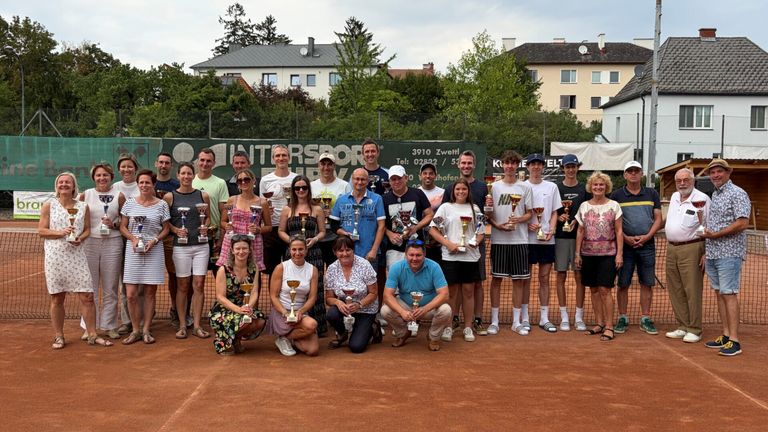 Eine Gruppe von Menschen mit Pokalen auf einem Tennisplatz, mit Gebäuden und Bäumen im Hintergrund.