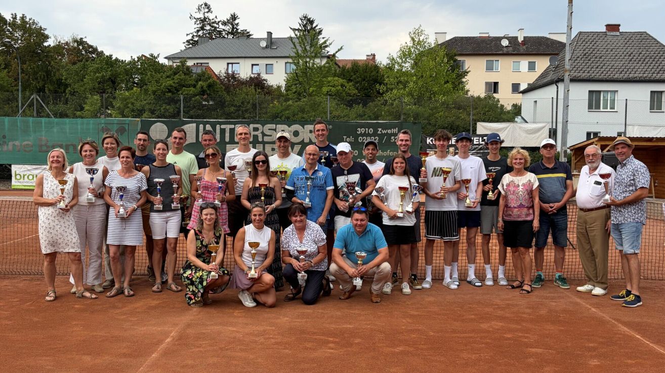 Eine Gruppe von Menschen mit Pokalen auf einem Tennisplatz, mit Gebäuden und Bäumen im Hintergrund.