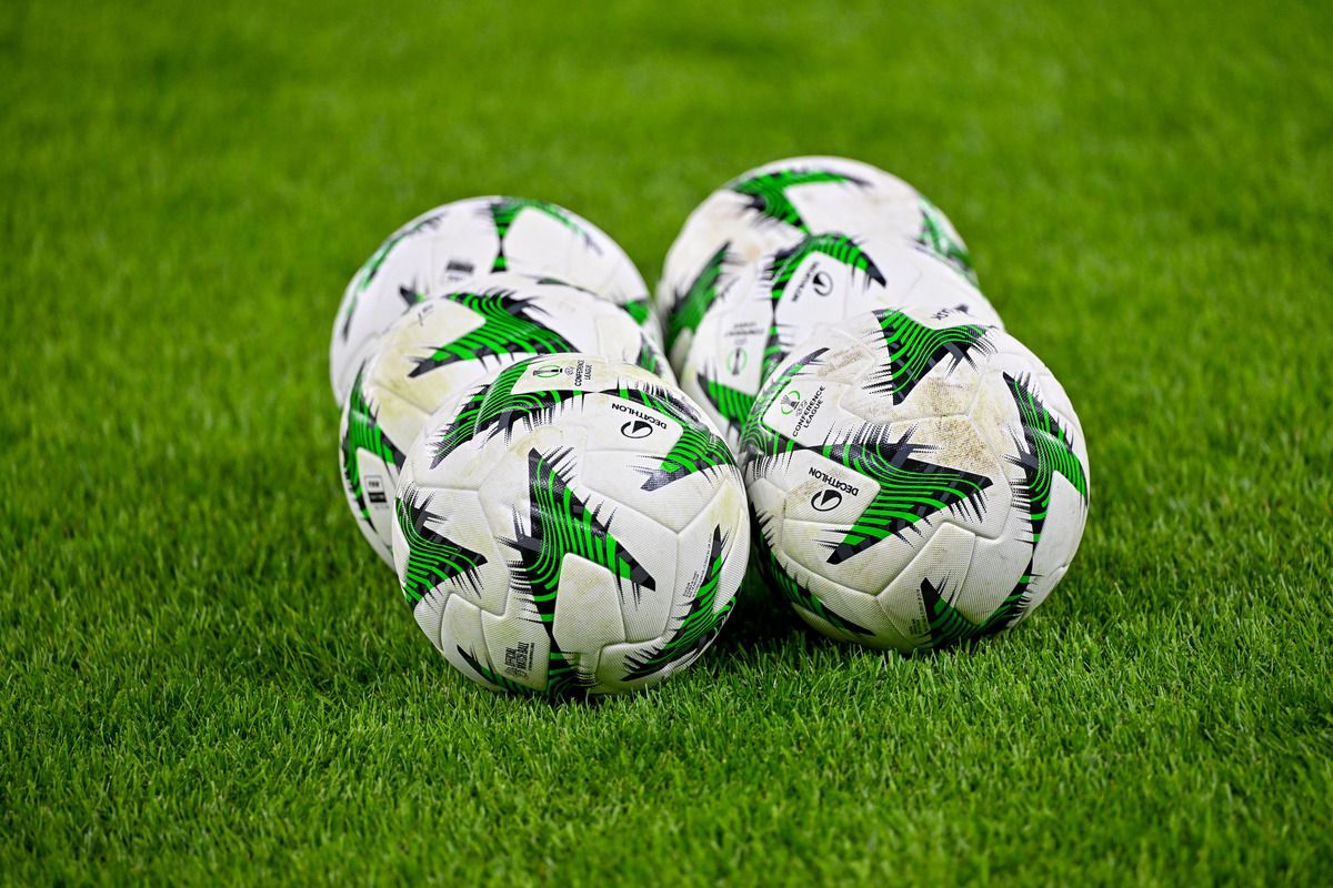 Fünf Fußballbälle mit weißen und grünen Designs liegen auf einem grünen Rasen. Die Bälle haben einen schwarzen Umriss und einige Schmutzflecken.
