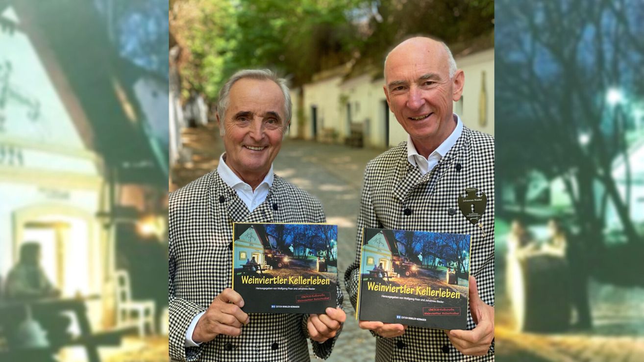 Zwei Männer in karierten Mänteln halten Bücher mit dem Titel 'Weinviertler Kellerleben' hoch und lächeln vor einem Gebäude mit Bäumen und einem Kopfsteinpflasterweg.
