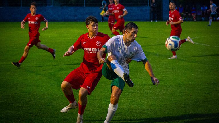 Zwei Fußballspieler in roten und weißen Trikots sind in einem Zweikampf auf einem Feld bei Nacht.