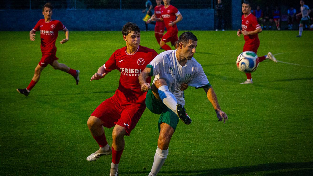 Zwei Fußballspieler in roten und weißen Trikots sind in einem Zweikampf auf einem Feld bei Nacht.