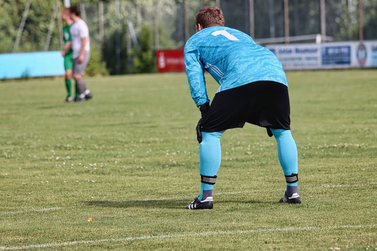 Ein Fußballspieler in einem blauen Trikot mit der Nummer 1 auf dem Rücken beugt sich auf einem Feld. Im Hintergrund stehen zwei Spieler in grünen und weißen Trikots.