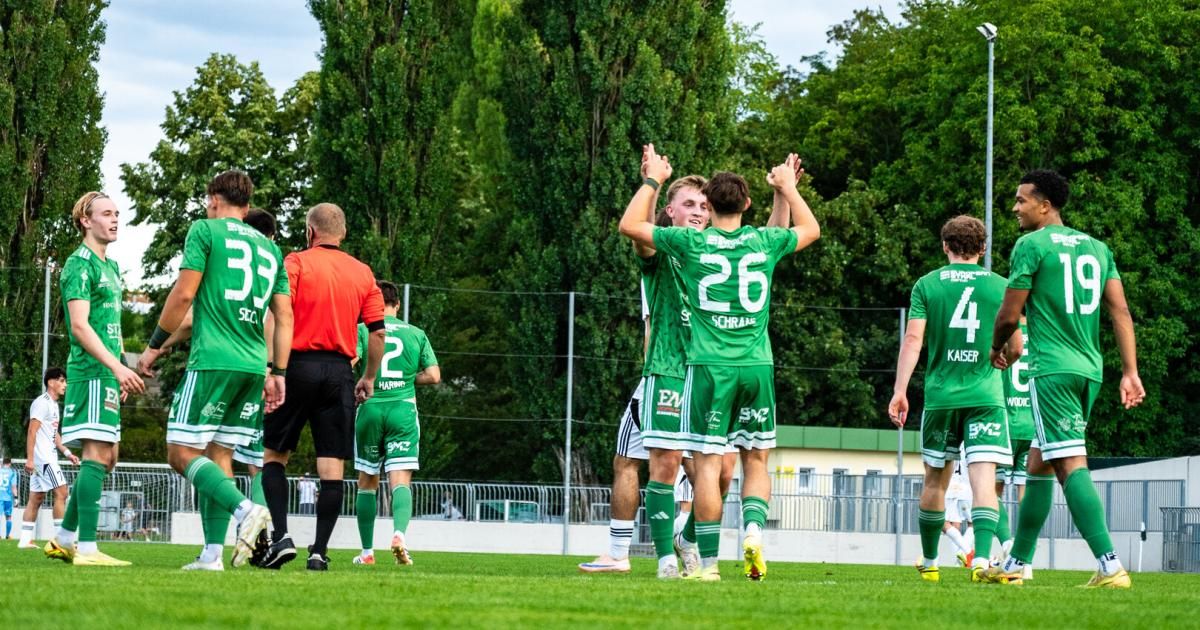 Fußballspieler in grünen Trikots feiern ein Tor auf einem Feld mit Bäumen im Hintergrund. Der Spieler in der Mitte hat die Nummer 26 auf seinem Trikot.