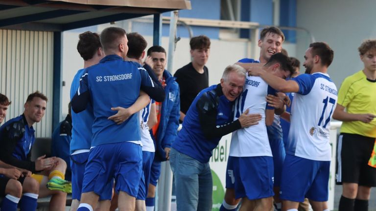 Eine Gruppe von Fußballspielern in blauen Trikots feiert mit einem Trainer, der eine blaue Jacke und Jeans trägt. Die Spieler umarmen sich und lächeln.