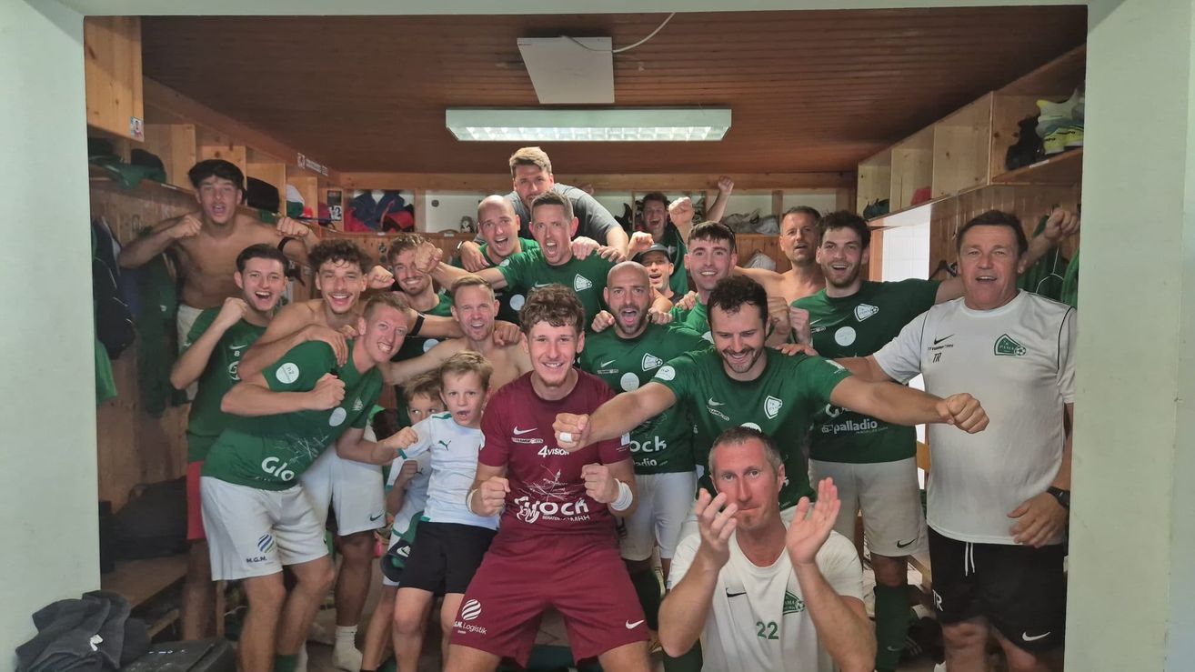 Eine Gruppe von Fußballspielern in grünen und burgunderroten Trikots posiert für ein Foto in einer Umkleidekabine. Die Spieler lächeln und machen verschiedene Gesten. Der Raum hat Holzwände und eine Decke mit Lichtern.