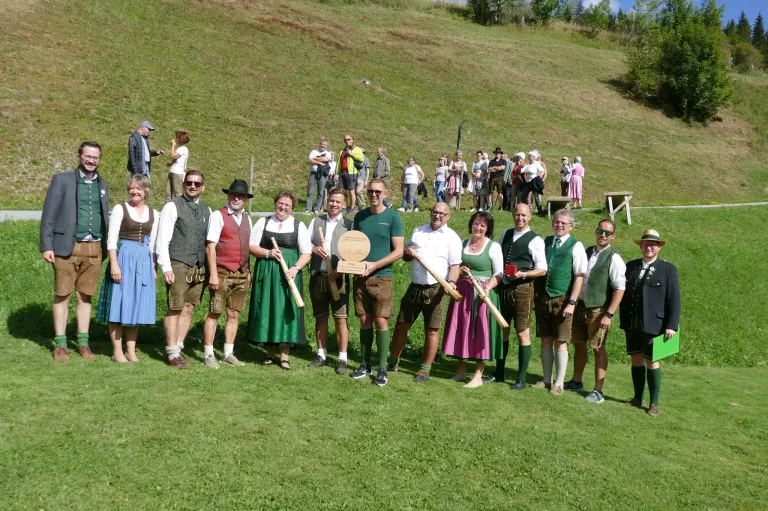 Eine Gruppe von Menschen in traditioneller Kleidung, die Holzstäbe halten und für ein Foto auf einem Grashügel mit einer Menschenmenge im Hintergrund posieren.