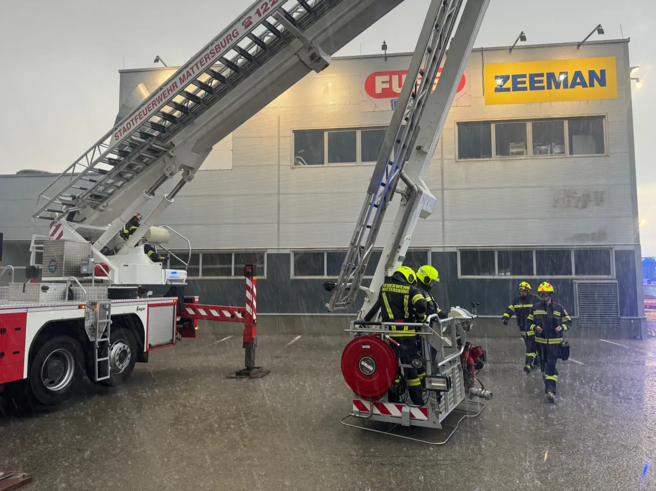 Ein Feuerwehrauto mit Feuerwehrleuten in gelben Helmen und Uniformen steht im Regen vor einem Gebäude mit einem Zeeman-Schild.