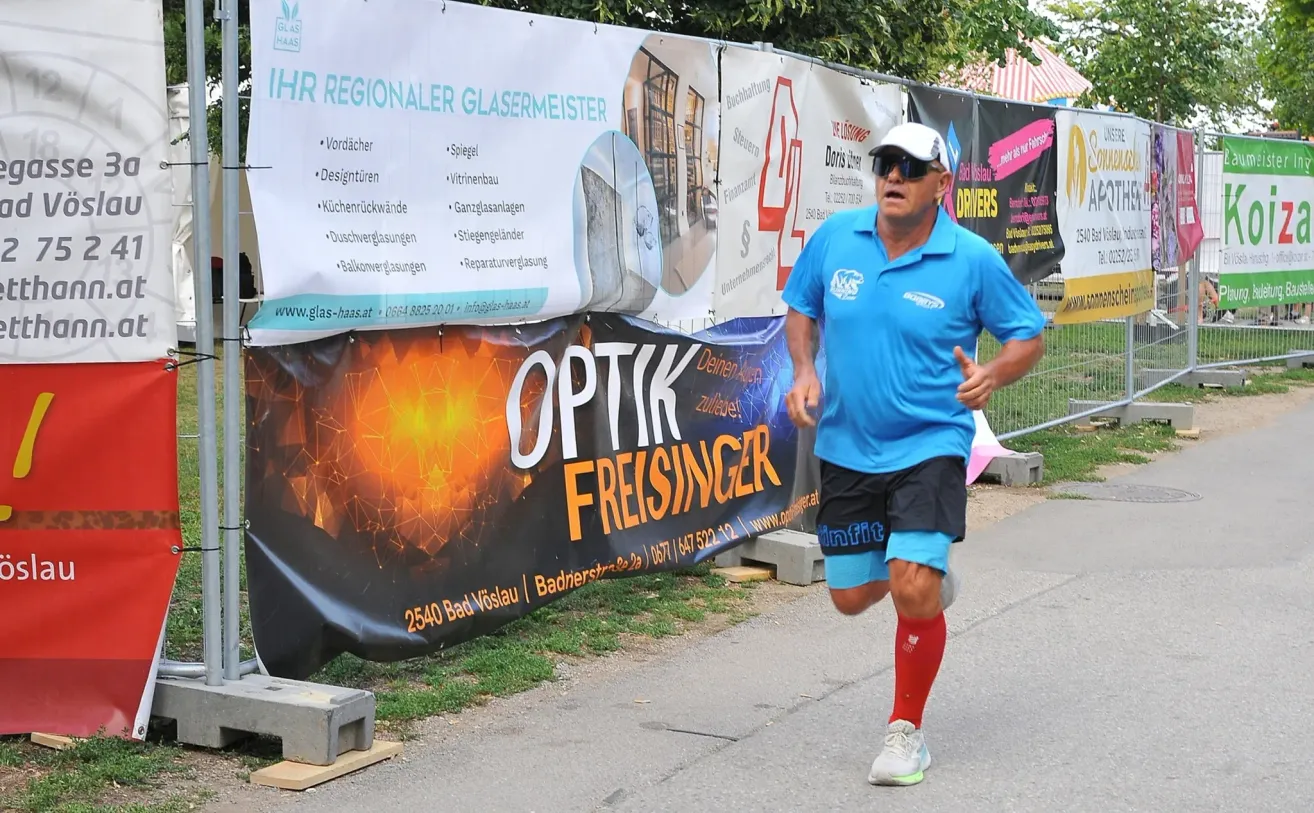 Ein Mann läuft an einer Straße mit einem Banner hinter ihm, das 'OPTIK FREISINGER' sagt. Er trägt ein blaues Shirt, schwarze Shorts und rote Socken.
