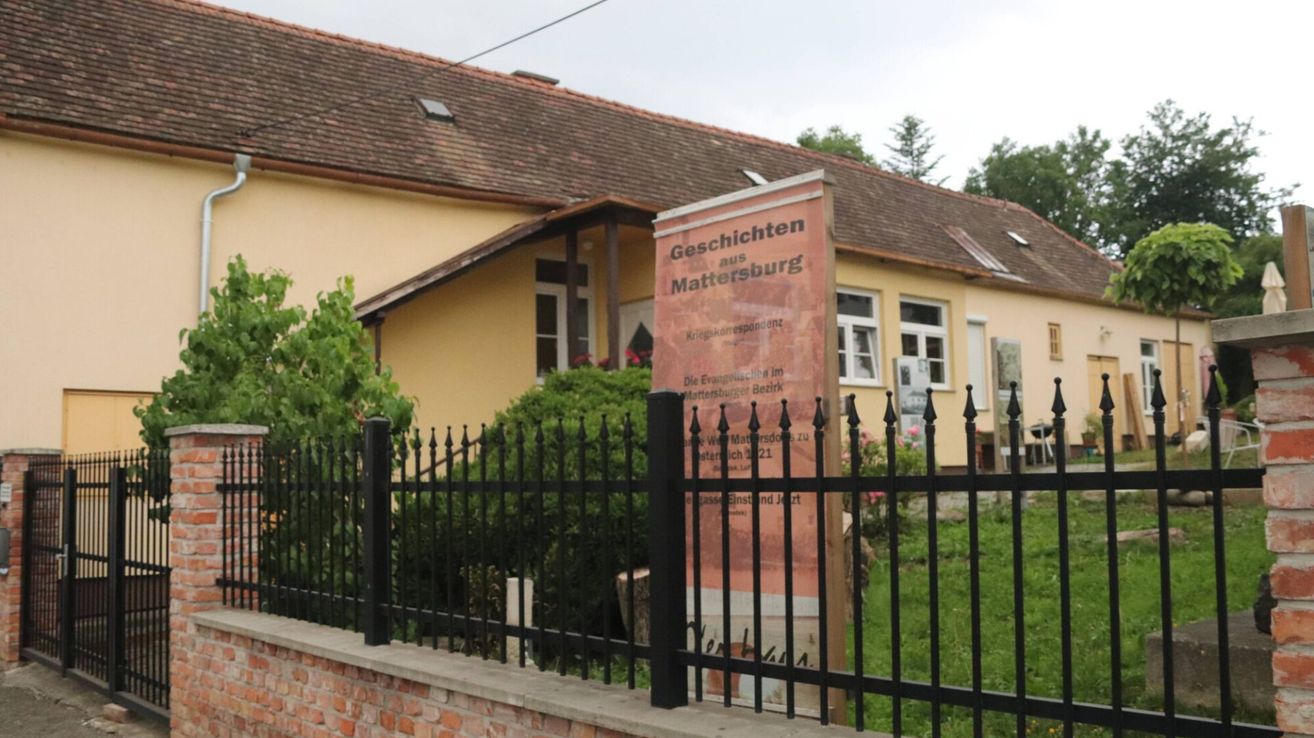 Ein Haus mit einem braunen Dach und gelben Wänden, mit einem Schild mit deutschem Text und einem schwarzen Zaun davor.