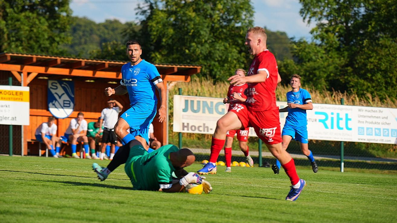 Ein Fußballspieler in Blau läuft auf das Tor zu, während ein anderer Spieler in Rot zu Boden fällt. Dahinter versucht ein Spieler in Grün, den Schuss zu blockieren.