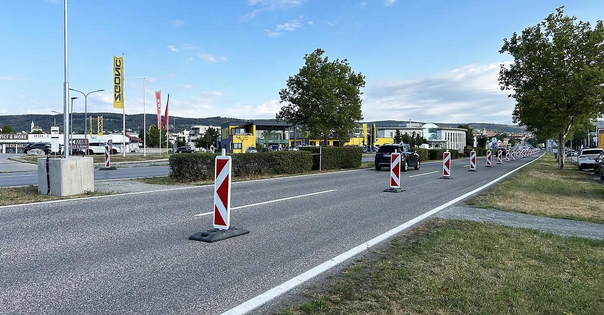 Eine Straße mit roten und weißen Verkehrskegel an der Seite, einem Baum und einem Gebäude in der Ferne.