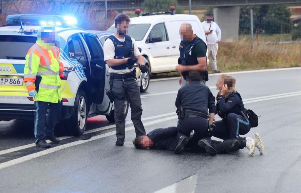 Polizisten nehmen einen Mann auf der Straße fest, während andere in der Nähe stehen. Ein Polizeiwagen und ein Auto sind dahinter geparkt.