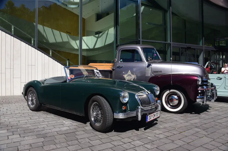 Ein klassischer grüner MG-Cabriolet ist vor einem modernen Gebäude geparkt, neben einem alten Polizeiwagen.