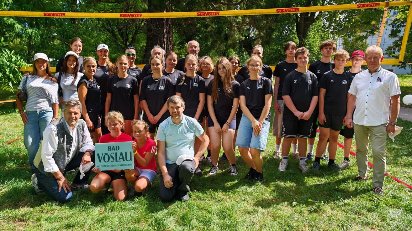 Eine Gruppe junger Menschen und Erwachsener in Sportkleidung posiert für ein Foto vor einem Volleyballnetz. Der Mann in der Mitte hält ein Schild mit der Aufschrift 'BAD VOSLA'.