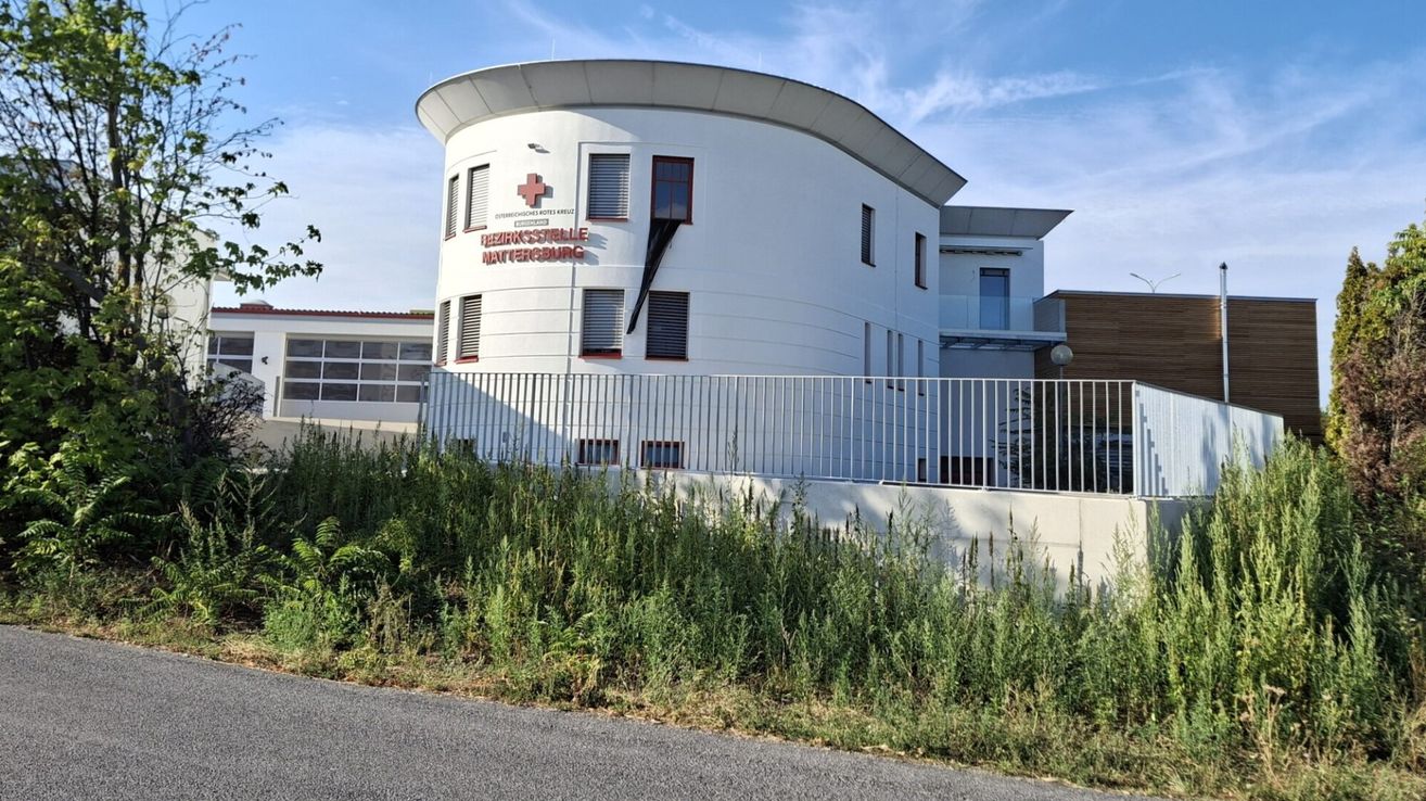 Ein modernes weißes Gebäude mit einem roten Kreuzschild auf der Fassade, benannt 'Rehaklinikstelle Mattersdorf', umgeben von einem Metallzaun und Grünpflanzen.