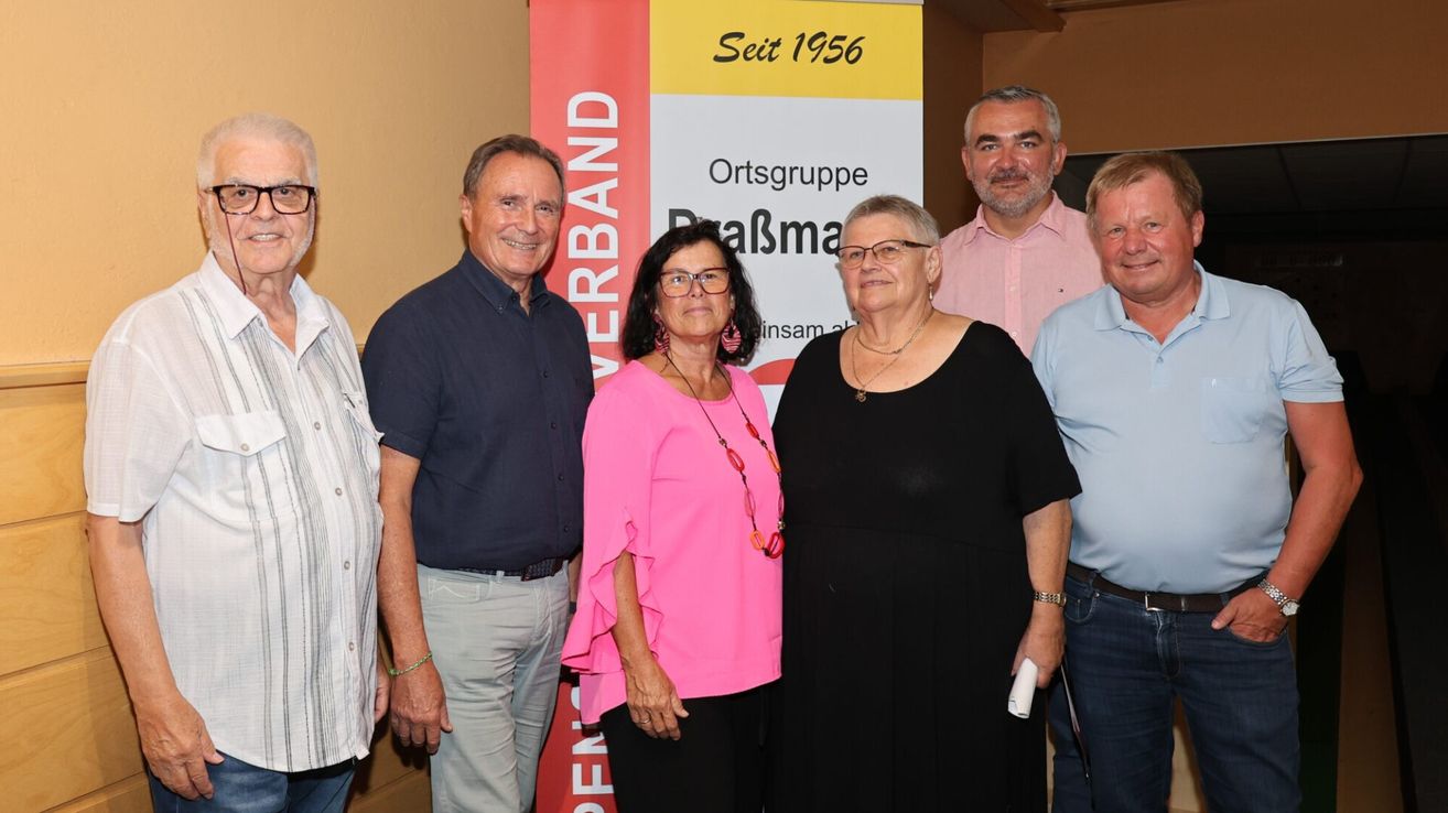 Fünf Personen stehen vor einem Banner, das 'Ortsgruppe Pralsma Seit 1956' besagt. Die Gruppe besteht aus zwei Frauen und drei Männern. Die Frau links trägt ein rosa Oberteil, die Frau in der Mitte ein schwarzes Kleid und der Mann rechts ein blaues Hemd.