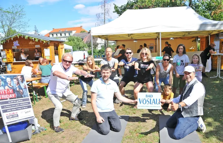 Eine Gruppe von Menschen macht Yoga im Freien. Einige von ihnen tragen Brillen und Turnschuhe. Ein Mann kniet auf dem Boden und hält ein Schild mit der Aufschrift 'BAD VOSLA'. Im Hintergrund befinden sich Zelte und ein Gebäude.