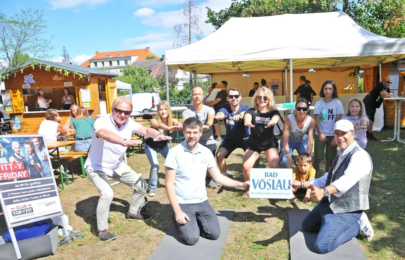 Eine Gruppe von Menschen macht Yoga im Freien. Einige von ihnen tragen Brillen und Turnschuhe. Ein Mann kniet auf dem Boden und hält ein Schild mit der Aufschrift 'BAD VOSLA'. Im Hintergrund befinden sich Zelte und ein Gebäude.