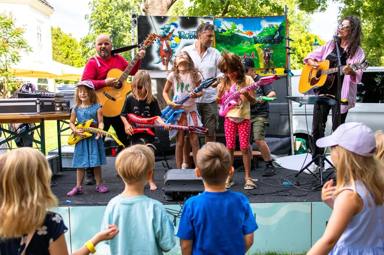 Eine Gruppe von Kindern und Erwachsenen spielt auf der Bühne Gitarre. Die Kinder halten pinke und gelbe Gitarren. Die Erwachsenen spielen Gitarre und stehen hinter den Kindern. Es gibt Lautsprecher und ein Mikrofon auf der Bühne. Im Hintergrund sind Bäume und ein Zelt zu sehen.