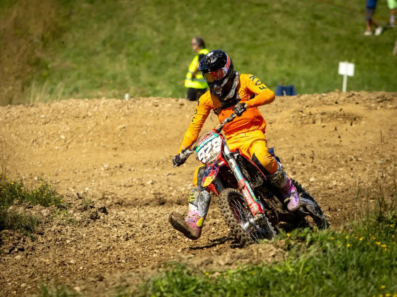 A motocross rider in full gear, riding on a dirt track, with a grassy background and a safety officer in the distance.