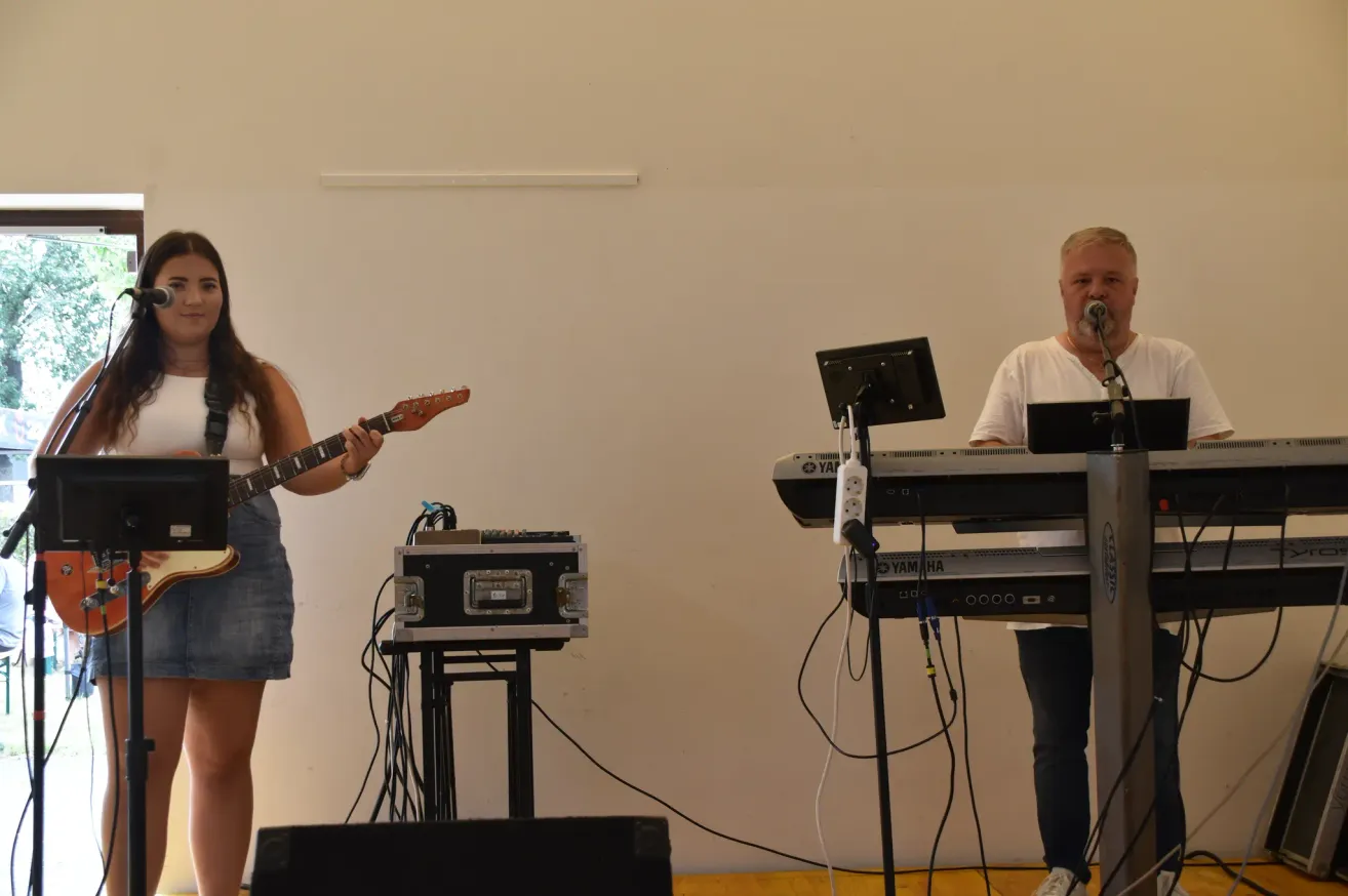 Ein Mann singt in ein Mikrofon, während eine Frau neben einem Keyboard auf der Bühne Gitarre spielt.