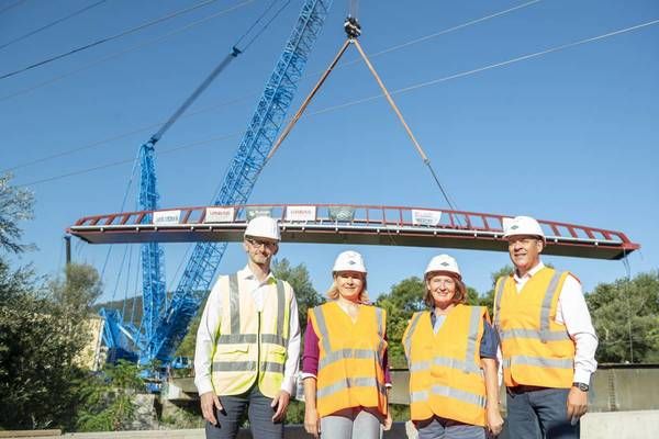 Vier Personen in Warnwesten stehen vor einer teilweise gebauten Brücke, mit einem großen blauen Kran im Hintergrund.