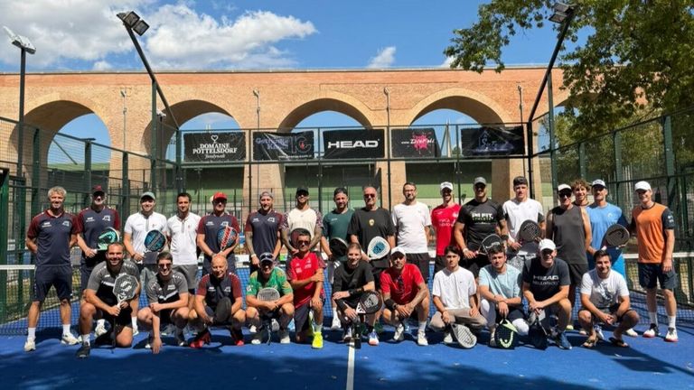 Eine Gruppe von Männern posiert für ein Foto auf einem blauen Tennisplatz mit Schlägern, in verschiedenen farbigen Shirts und Mützen. Hinter ihnen steht ein Backsteingebäude mit Bogenfenstern und Bannern.