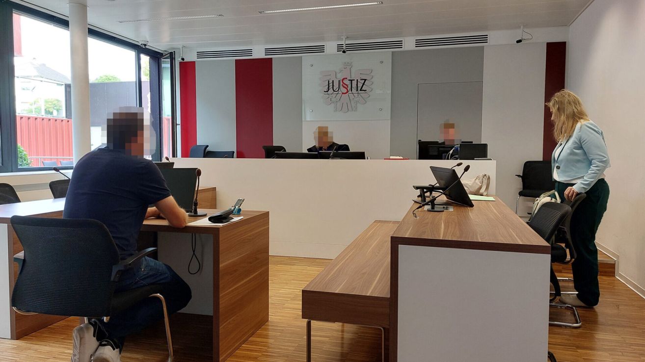 Ein Büro mit einem verwischten Menschen am Schreibtisch, einem 'Justiz'-Schild an der Wand und Monitoren und Mikrofonen auf dem Schreibtisch.