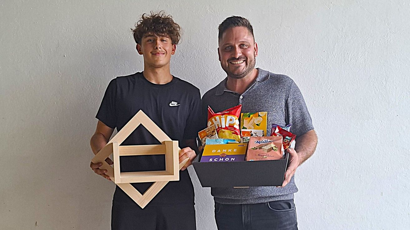 Two men stand next to each other, one holding a wooden box and the other holding a box of snacks. They both smile for the camera.
