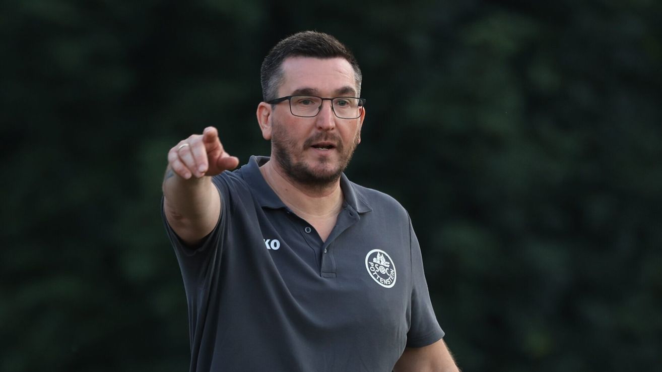 A man wearing a gray polo shirt with a white logo on the left chest area is pointing with his right hand. He is wearing glasses and has a beard.