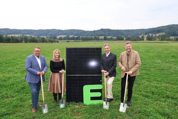 Vier Personen stehen in einem Feld mit Schaufeln, lächelnd, und posieren neben einem großen Solarpanel. Der Buchstabe E ist prominent auf dem Panel angebracht.