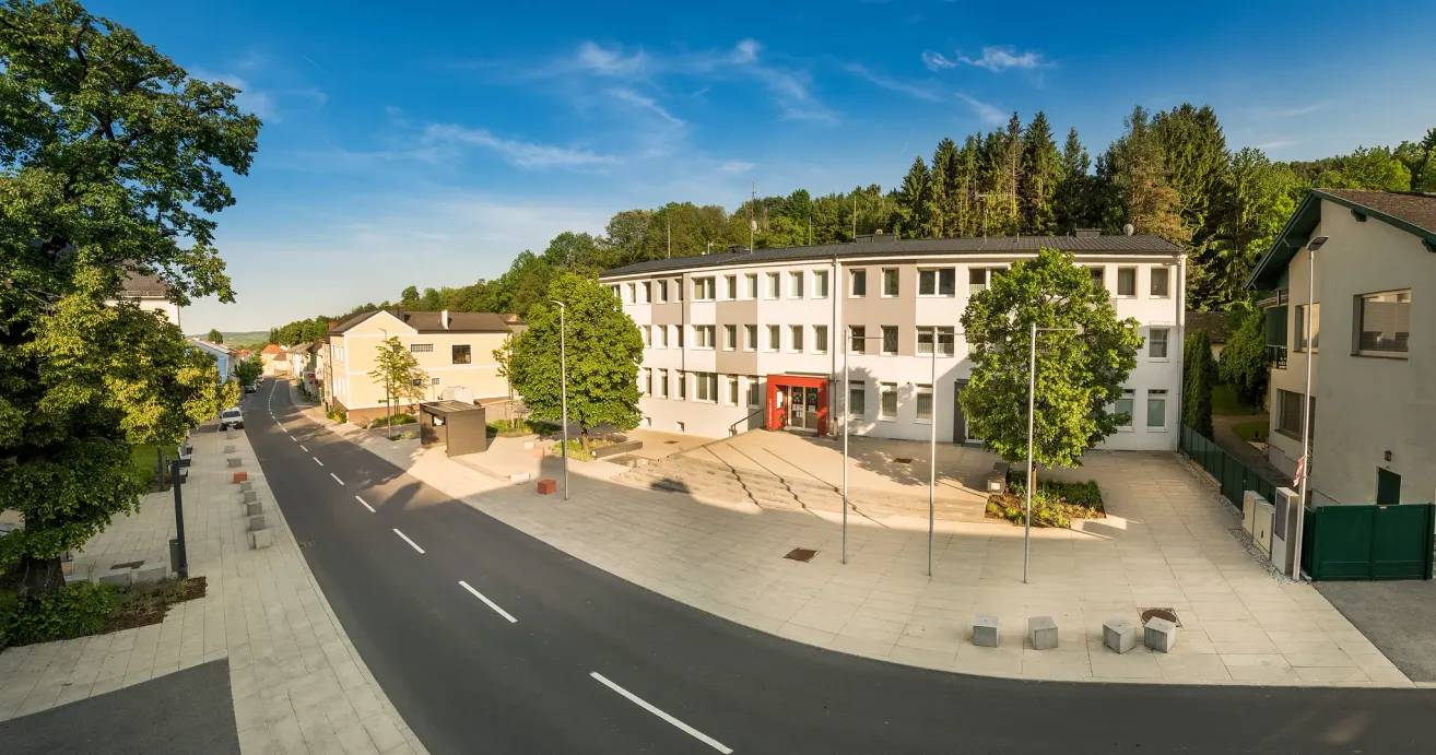 Eine Luftaufnahme eines modernen Gebäudes mit einem roten Eingang, umgeben von Bäumen und einem gepflasterten Gehweg. Das Gebäude steht vor einem Hintergrund eines klaren blauen Himmels mit einigen Wolken.