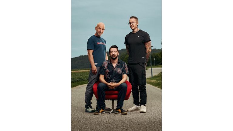 Drei Männer stehen auf einer Straße, einer sitzt auf einem roten Stuhl, ein anderer steht hinter ihm und der dritte steht neben ihnen. Sie sind lässig gekleidet und scheinen für ein Foto zu posieren.
