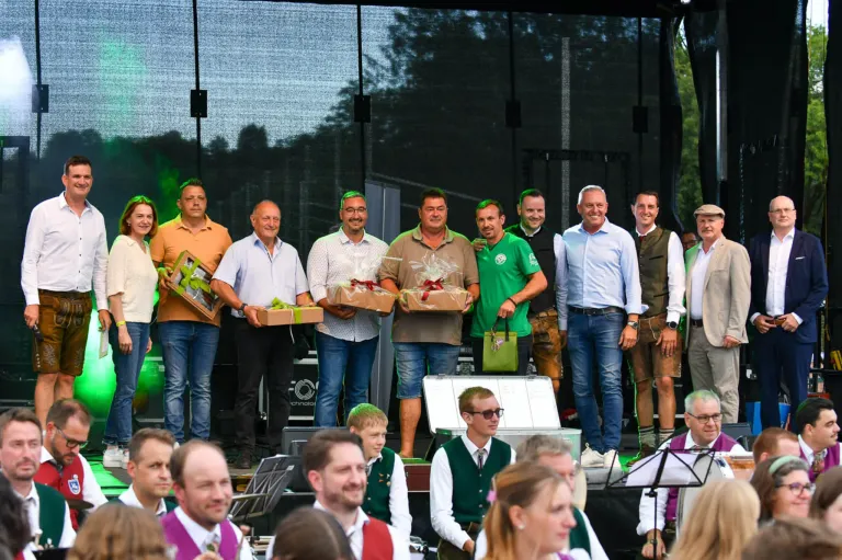 Eine Gruppe von Männern und Frauen steht auf einer Bühne und hält Geschenkboxen in der Hand, mit einer großen Leinwand und Bäumen im Hintergrund.