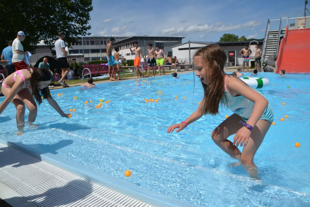 Ein kleines Mädchen läuft in einem Schwimmbecken mit schwimmenden gelben Bällen, während andere schwimmen und herumstehen.