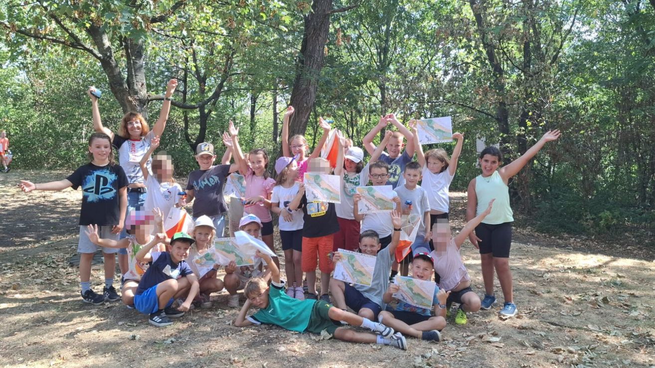 Eine Gruppe von Kindern im Wald, die Karten halten und für ein Foto posieren, einige sitzend und andere stehend, alle lächeln.