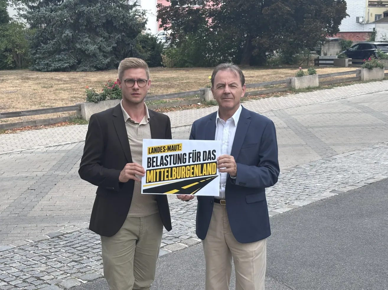 Zwei Männer stehen auf einer Kopfsteinpflasterstraße, einer hält ein Schild mit der Aufschrift 'Landes-Maut: Belastung für das Mittelburgenland'. Der Mann links trägt eine Brille und einen dunklen Blazer, während der Mann rechts einen blauen Blazer und eine Uhr trägt.