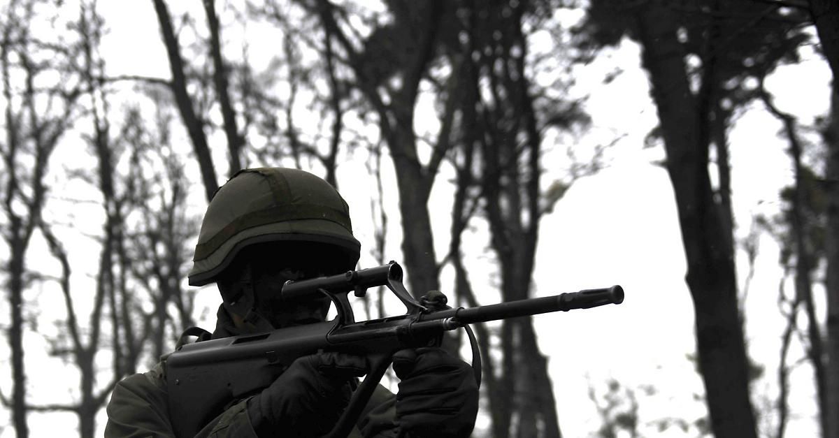 Ein Soldat in Helm und Handschuhen hält ein Gewehr in einem Wald.
