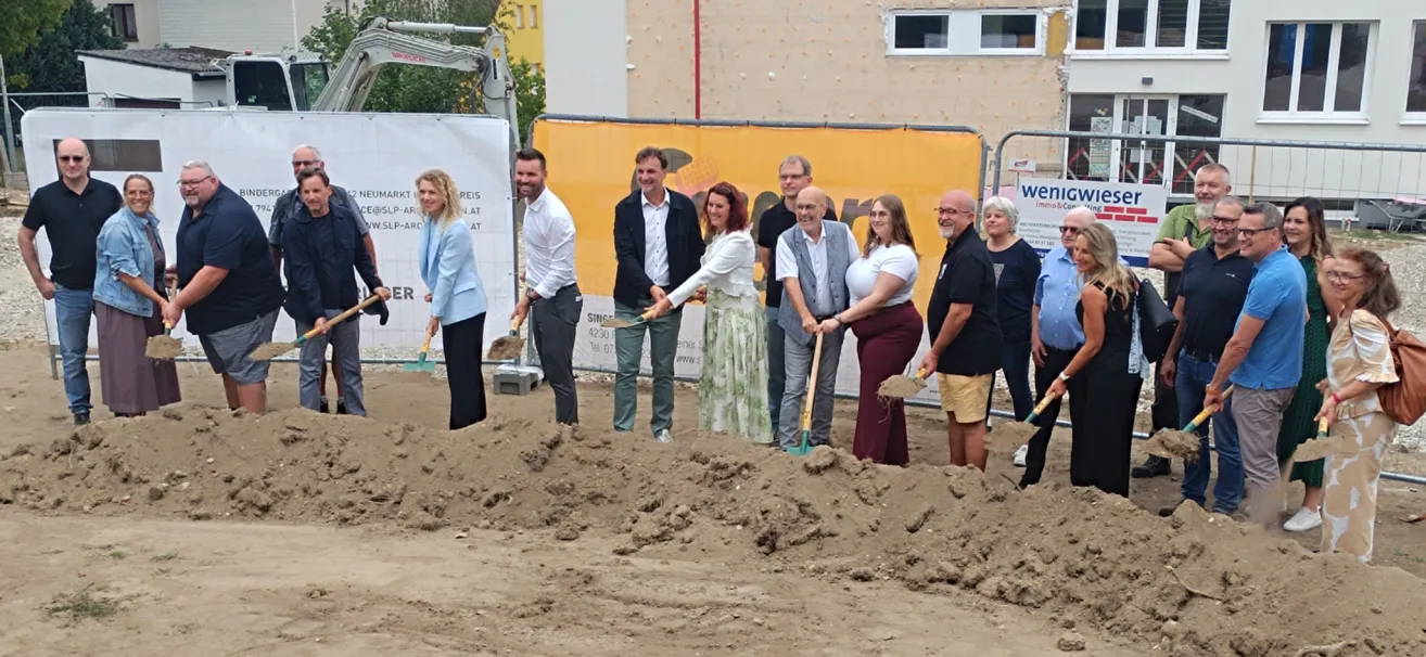 A group of people in formal attire are participating in a groundbreaking ceremony, holding shovels and standing in a pile of dirt. Behind them, a building under construction is visible.
