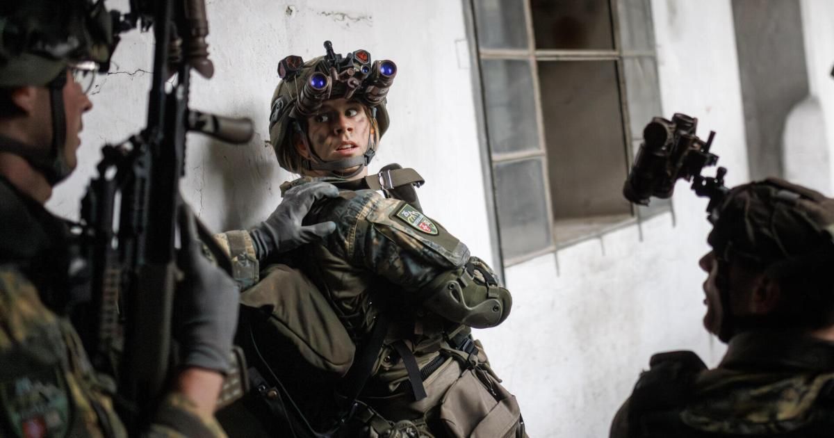 Ein Soldat mit Nachtsichtbrille und Tarnuniform wird von einem anderen Soldaten in einem Raum mit weißer Wand und offenem Fenster unterstützt.