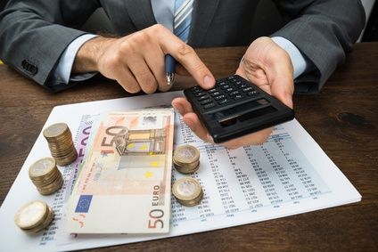 Ein Mann in Anzug berechnet mit einem Taschenrechner und hält einen Stapel Euro-Banknoten und Münzen auf einem Schreibtisch.