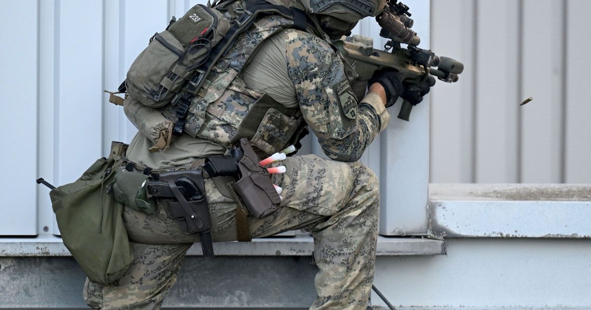 Ein Soldat in Tarnkleidung, ausgestattet mit einem Gewehr, einer Seitenwaffe und verschiedenen Taschen, kauert hinter einer Betonmauer.