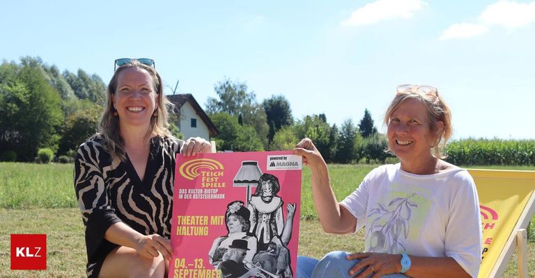 Zwei Frauen halten ein rosa Plakat mit einer Theaterveranstaltungsankündigung. Das Plakat zeigt eine Zeichnung einer Frau mit einer Lampe und Text auf Deutsch. Die Frauen lächeln und scheinen für ein Foto zu posieren. Dahinter befindet sich ein grasbewachsenes Feld mit Bäumen und einem Haus.