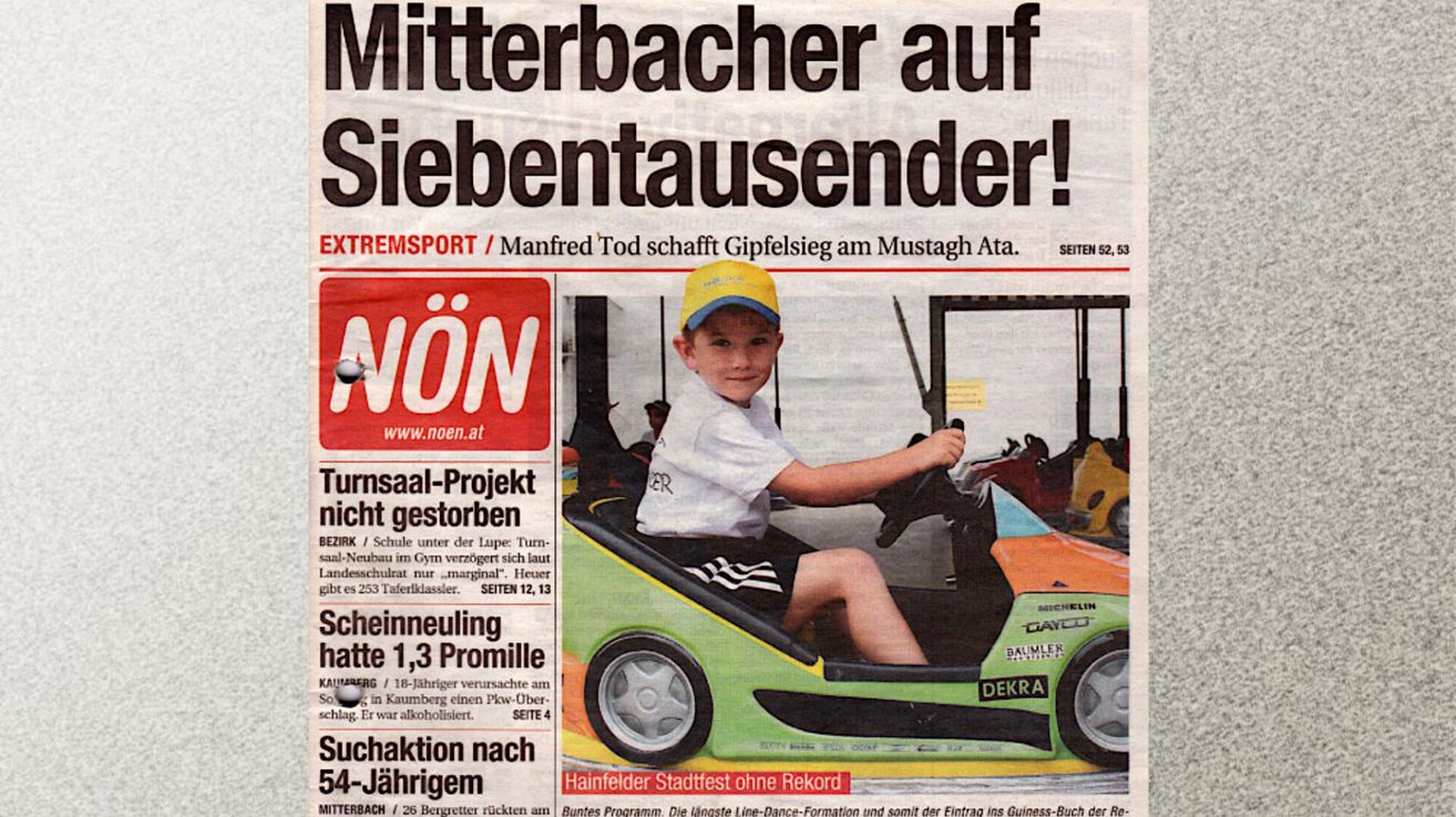 Ein Zeitungsausschnitt zeigt einen jungen Jungen, der einen bunten Go-Kart mit dem Wort 'DEKRA' an der Seite fährt. Die Schlagzeile lautet 'Mitterbacher auf Siebentausendsender!' und enthält weitere Nachrichtenartikel.