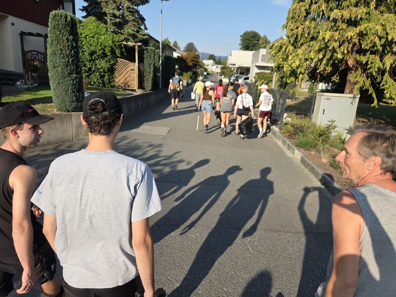 A group of people are walking down a street, some carrying bags and wearing hats. The shadows of the people are cast on the road.