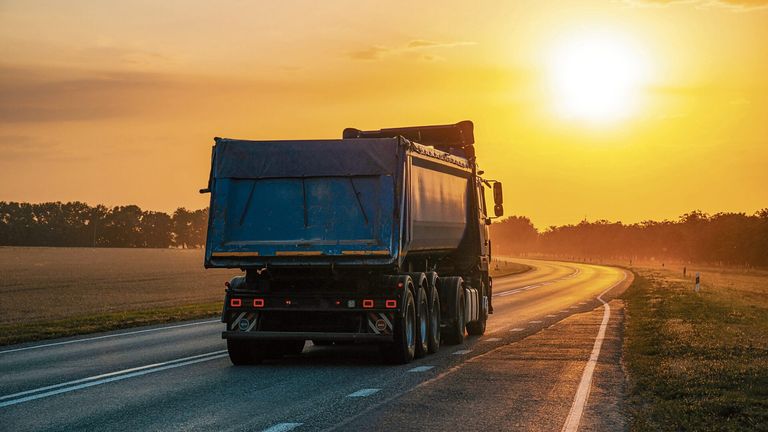 Ein blauer Kipplaster fährt bei Sonnenuntergang mit klarem Himmel und entfernten Bäumen auf einer Straße.