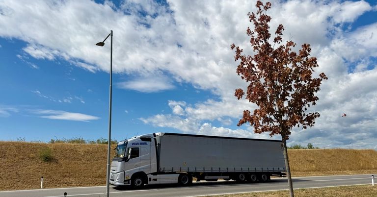 Ein weißer Volvo-Mietwagen fährt auf einer Straße mit einem bewölkten Himmel und einem Baum mit braunen Blättern.