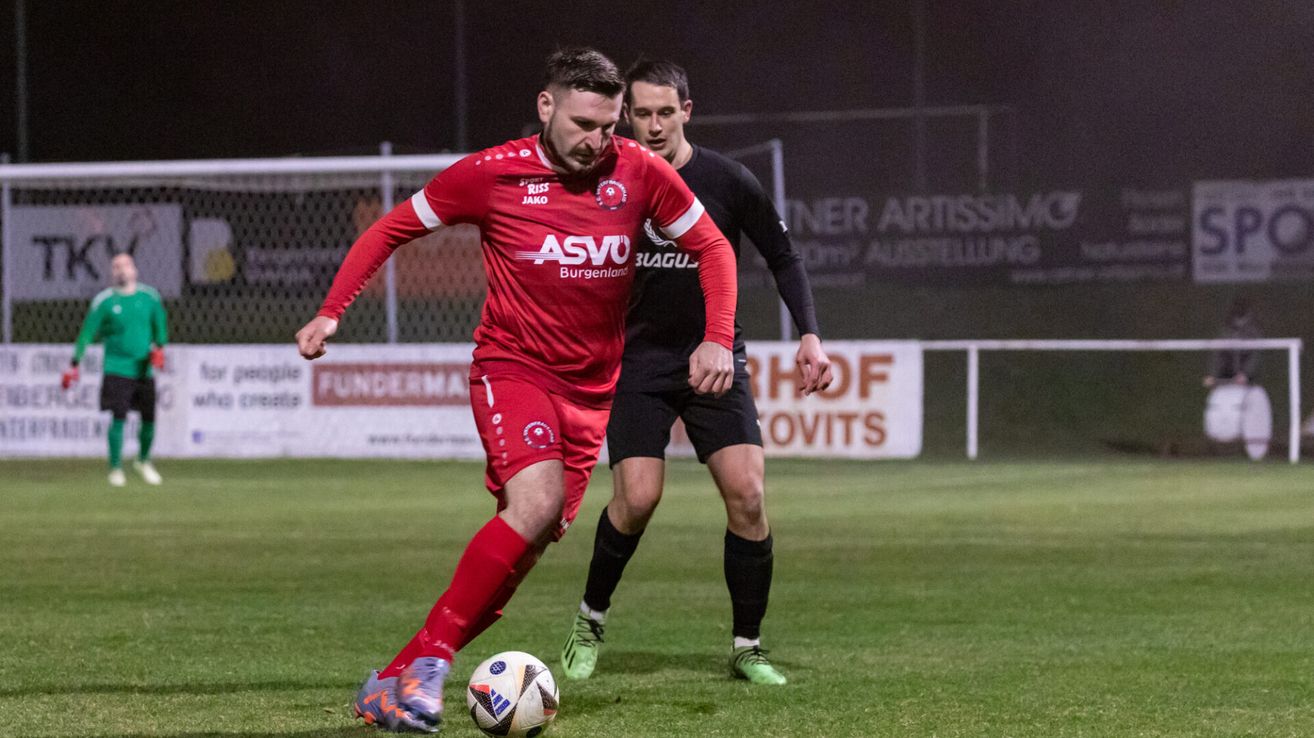 A soccer player in a red uniform is dribbling the ball while another player in black defends him on a soccer field at night.