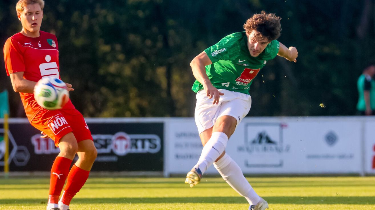 Ein Fußballspieler in einem grünen Trikot mit 'Synergo' darauf läuft und den Ball auf einem Feld tritt.